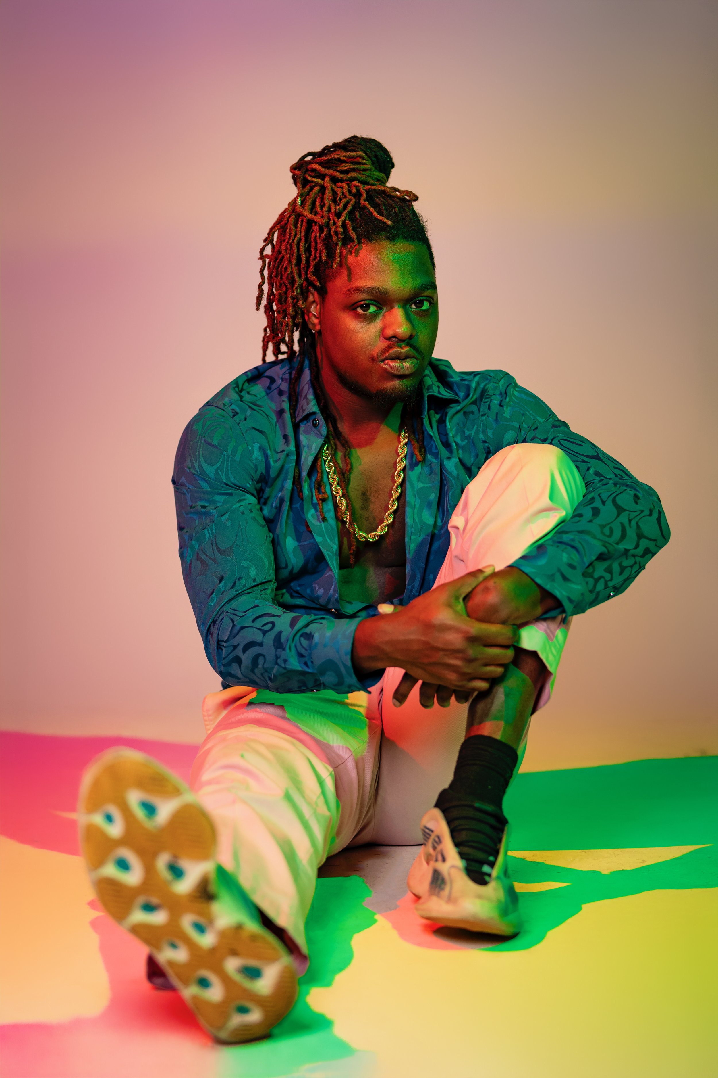 A young man with dreadlocks in a bun, wearing a blue patterned shirt, white pants, black socks, and athletic shoes, sitting on the floor with one knee up and holding his ankle, against a multicolored background with pink, green, and beige lighting.