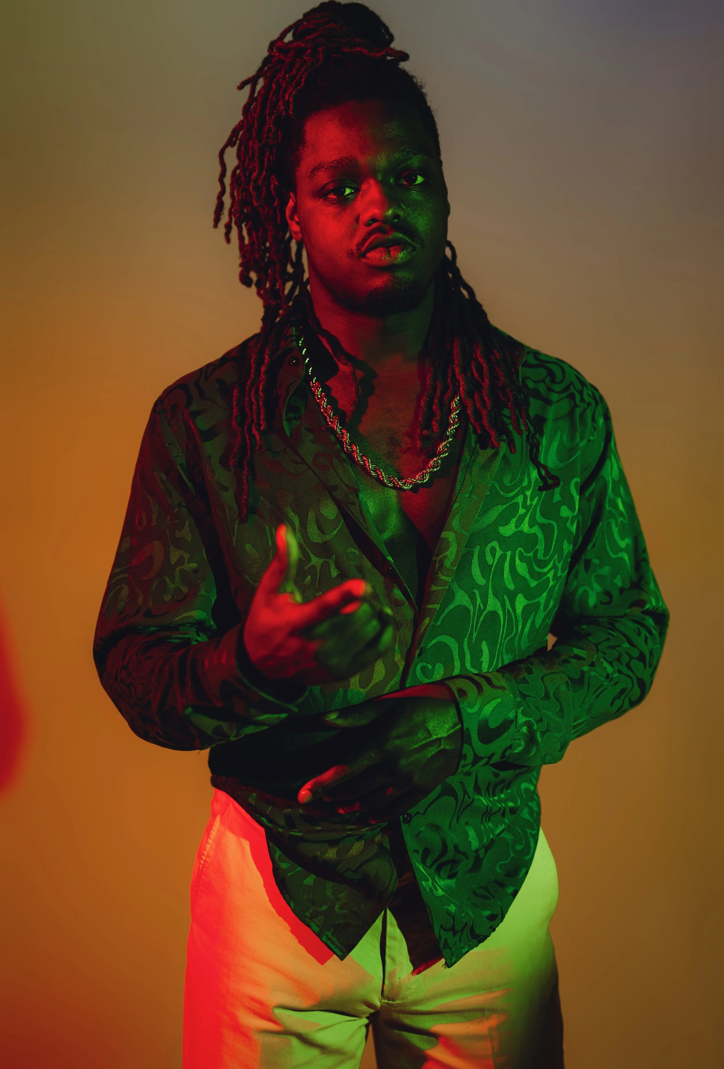 A man with long dreadlocks wearing a patterned shirt, layered necklaces, and beige pants, standing against a neutral background with colorful red and green lighting.