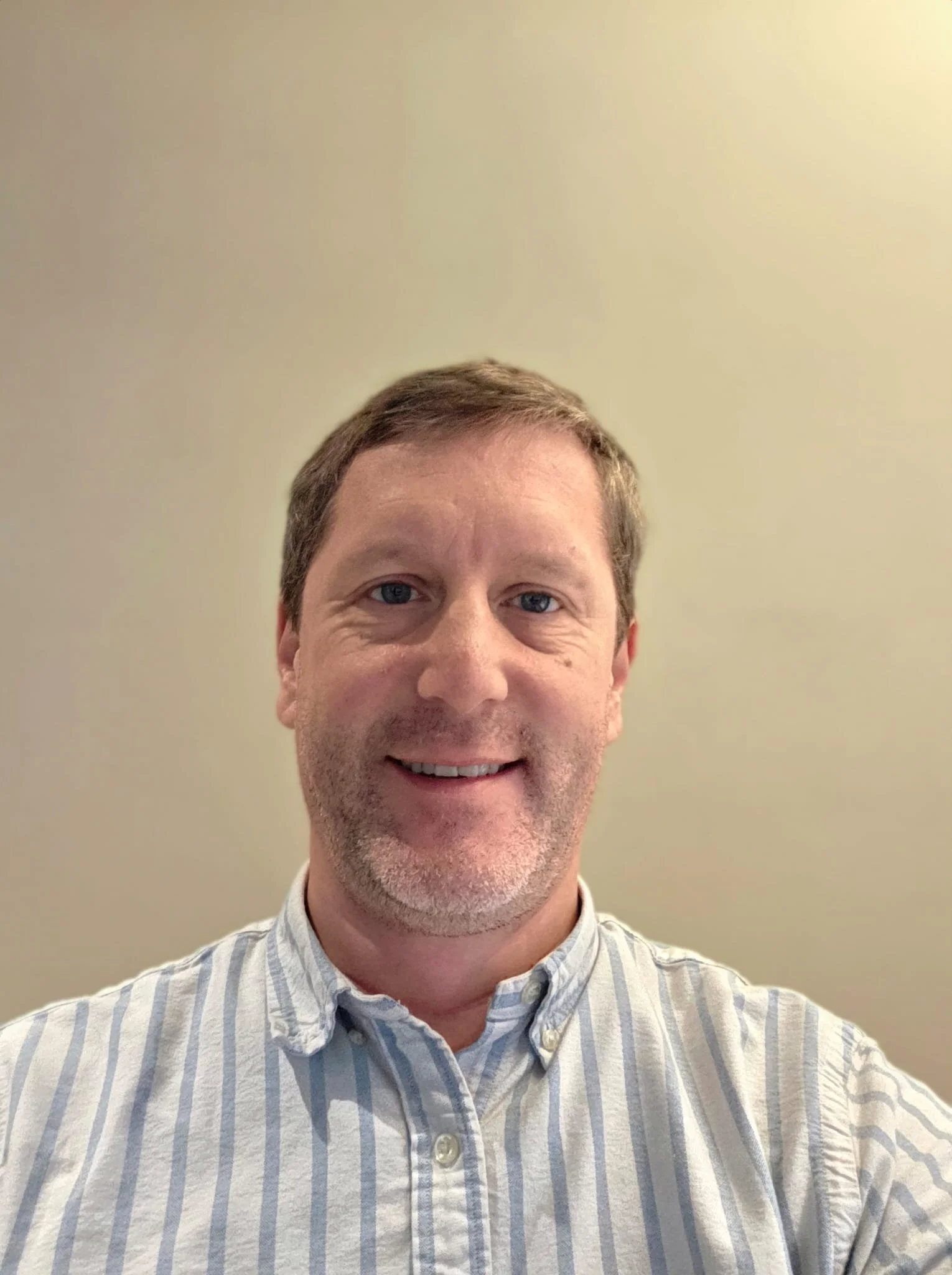 A middle-aged man with short brown hair and a light beard, wearing a collared, button-down shirt with vertical stripes, smiling in front of a plain beige wall.