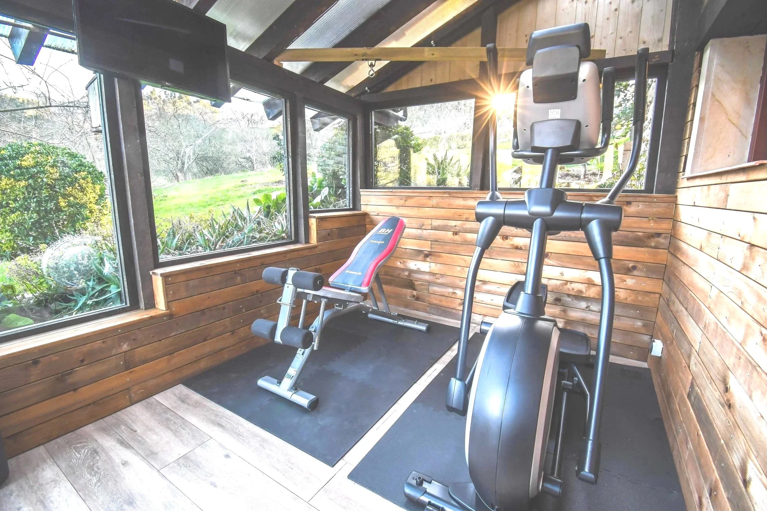 Gimnasio con máquina elíptica y banco de ejercicios en una habitación con paredes de madera y ventanas que dejan ver un jardín exterior.