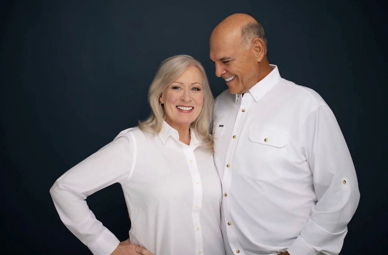 A smiling elderly couple wearing white shirts standing close together against a dark background.