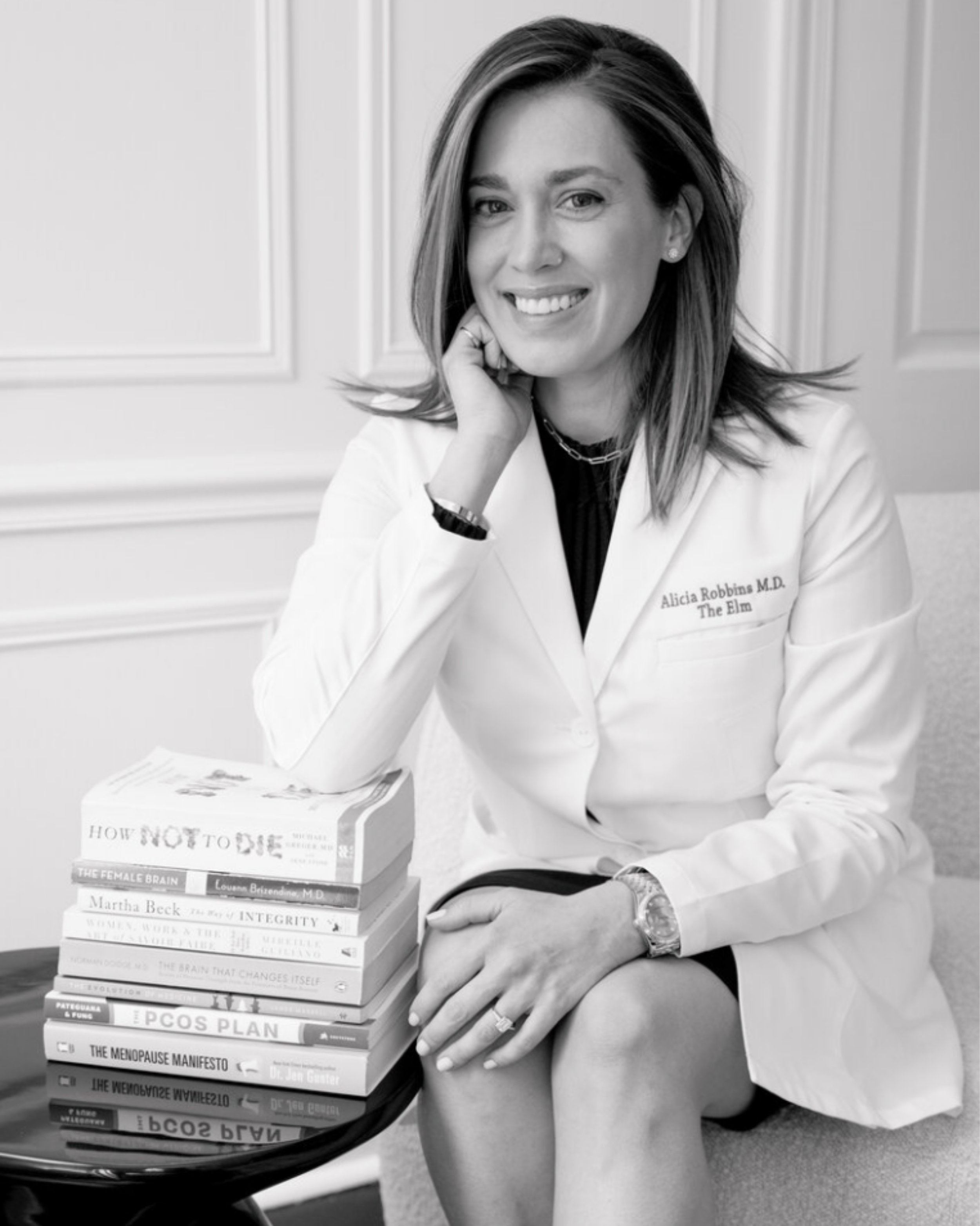 Black and white photo of a smiling woman in a white medical coat sitting next to a stack of health-related books.