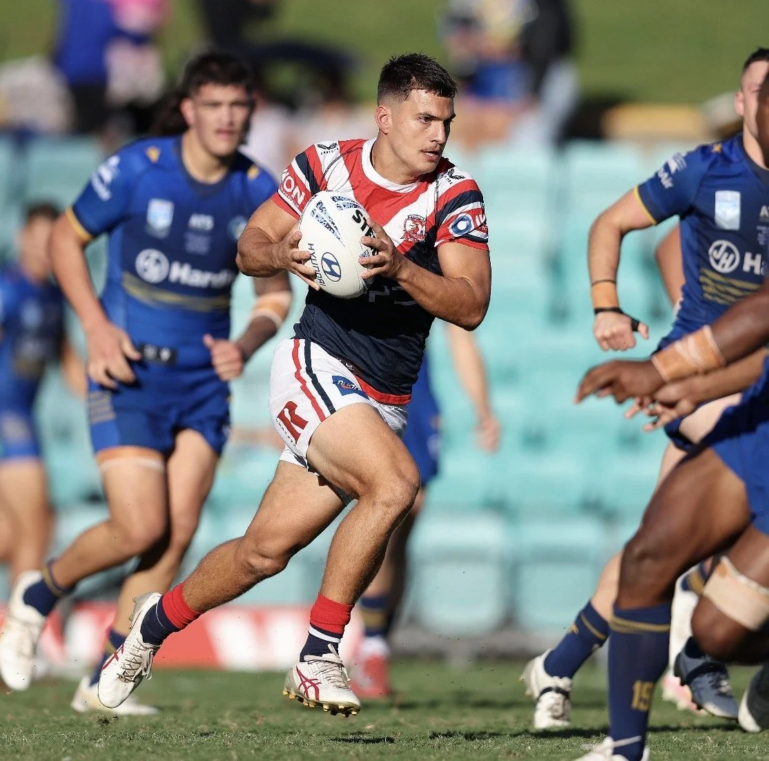 A rugby player in a red, white, and blue uniform holding a rugby ball while running during a match, with opponents in blue uniforms pursuing him.