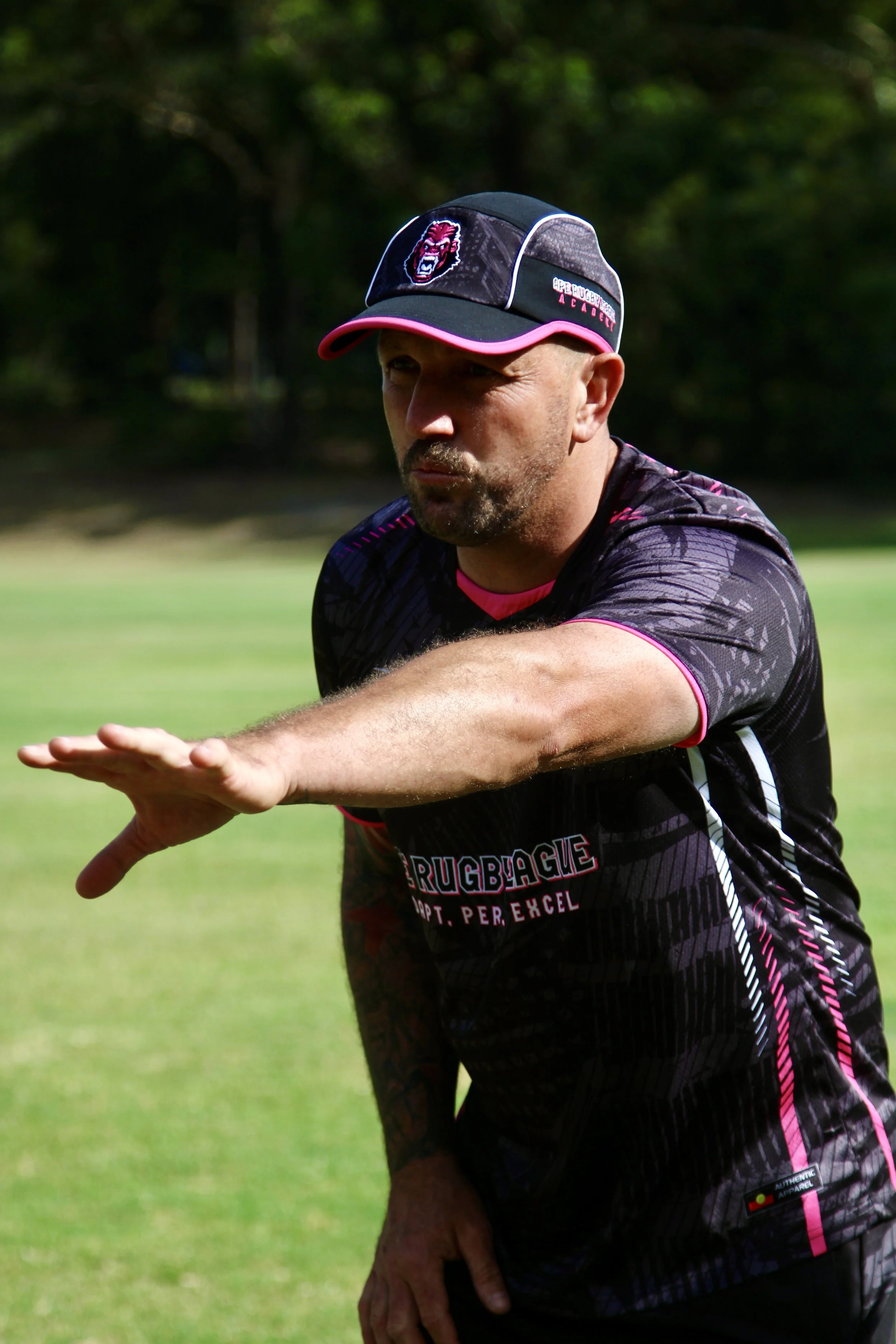 A man in sportswear and a cap extends his arm forward on a grassy field, possibly during a rugby training session.