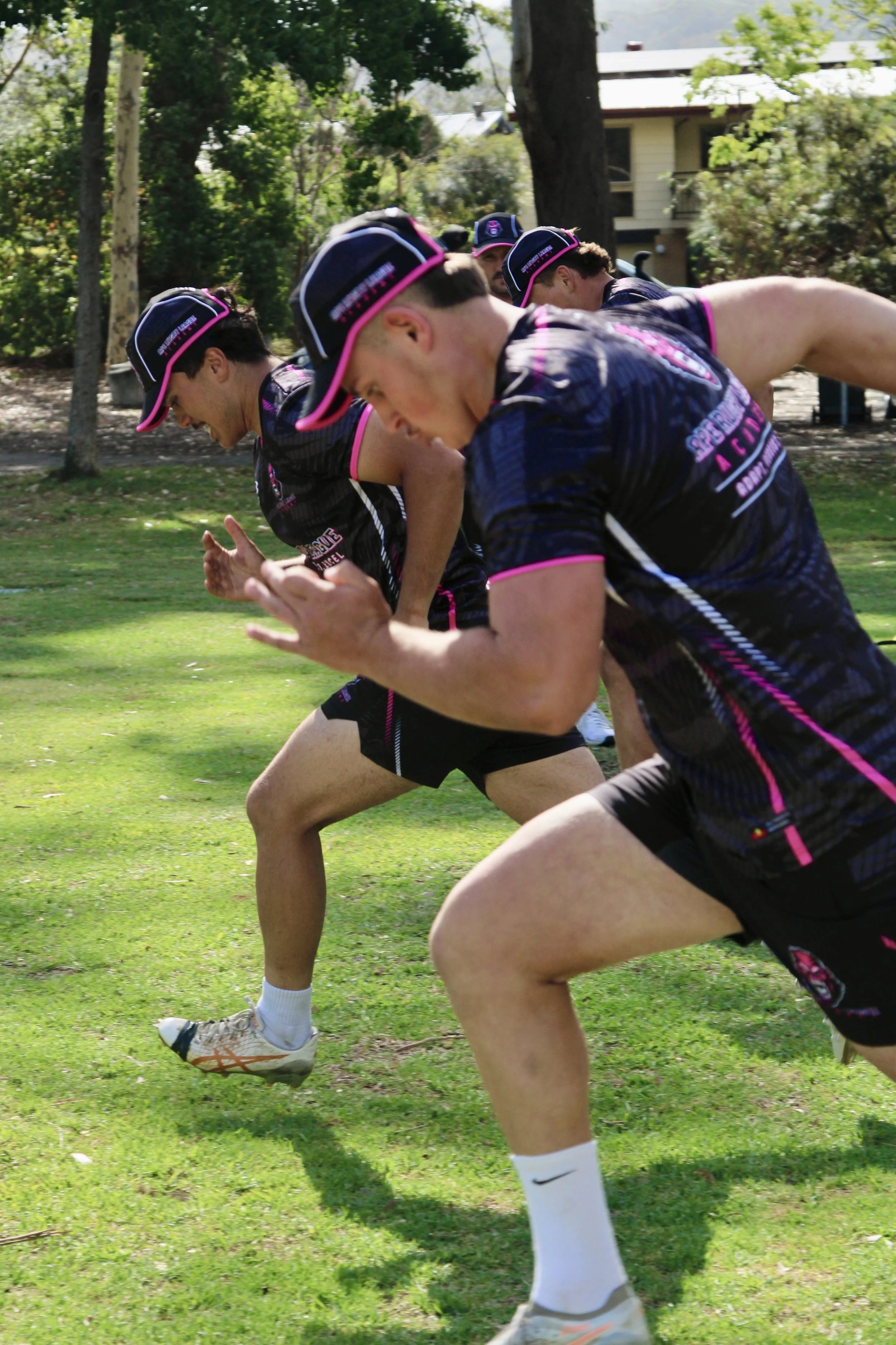 Group of young men running or sprinting outdoors on a grassy field during daytime, wearing matching black and pink athletic uniforms and caps.