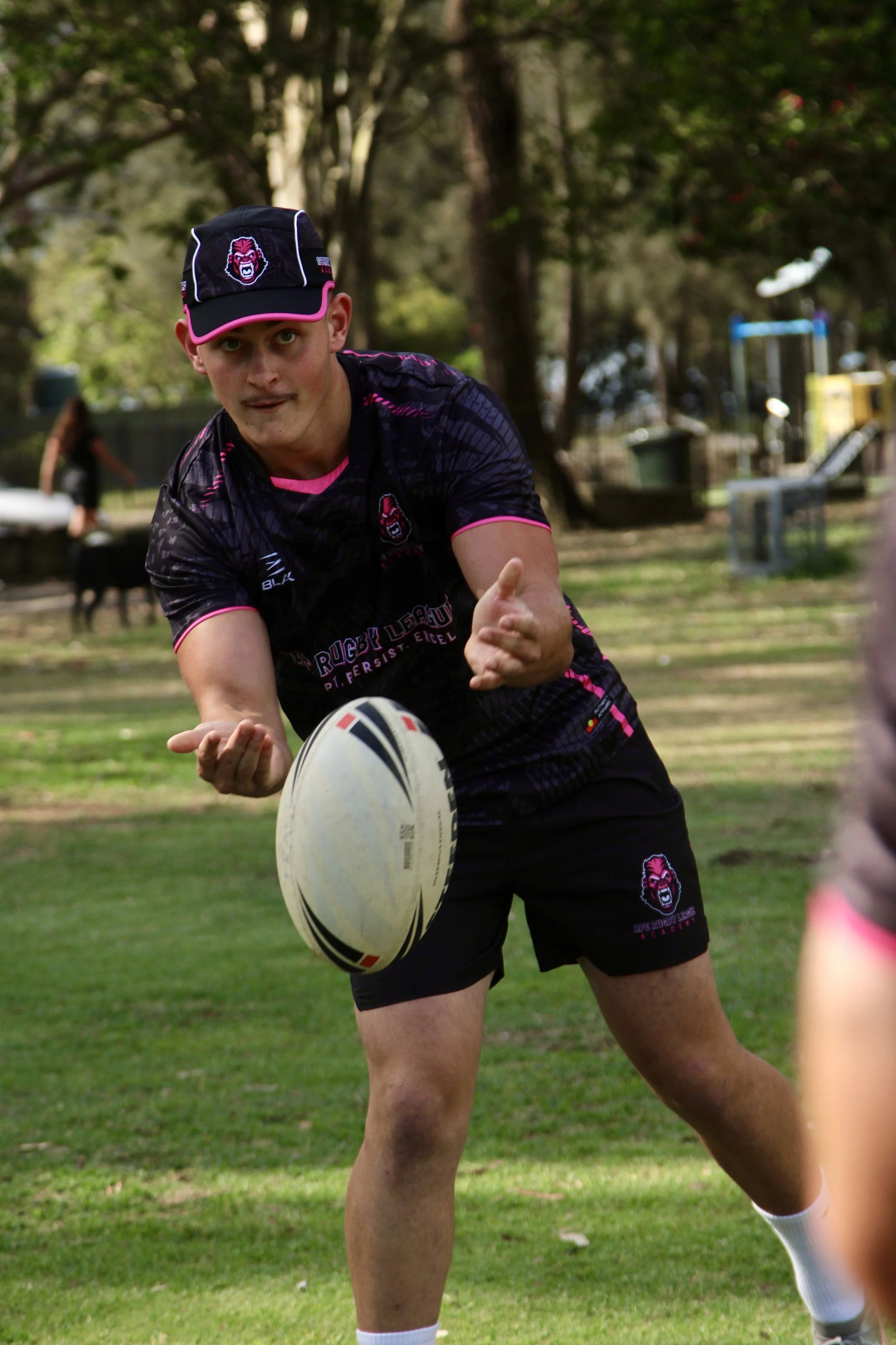 A young man playing rugby, reaching out to catch or pass a rugby ball in a park with trees in the background, wearing a black and pink rugby uniform and cap.