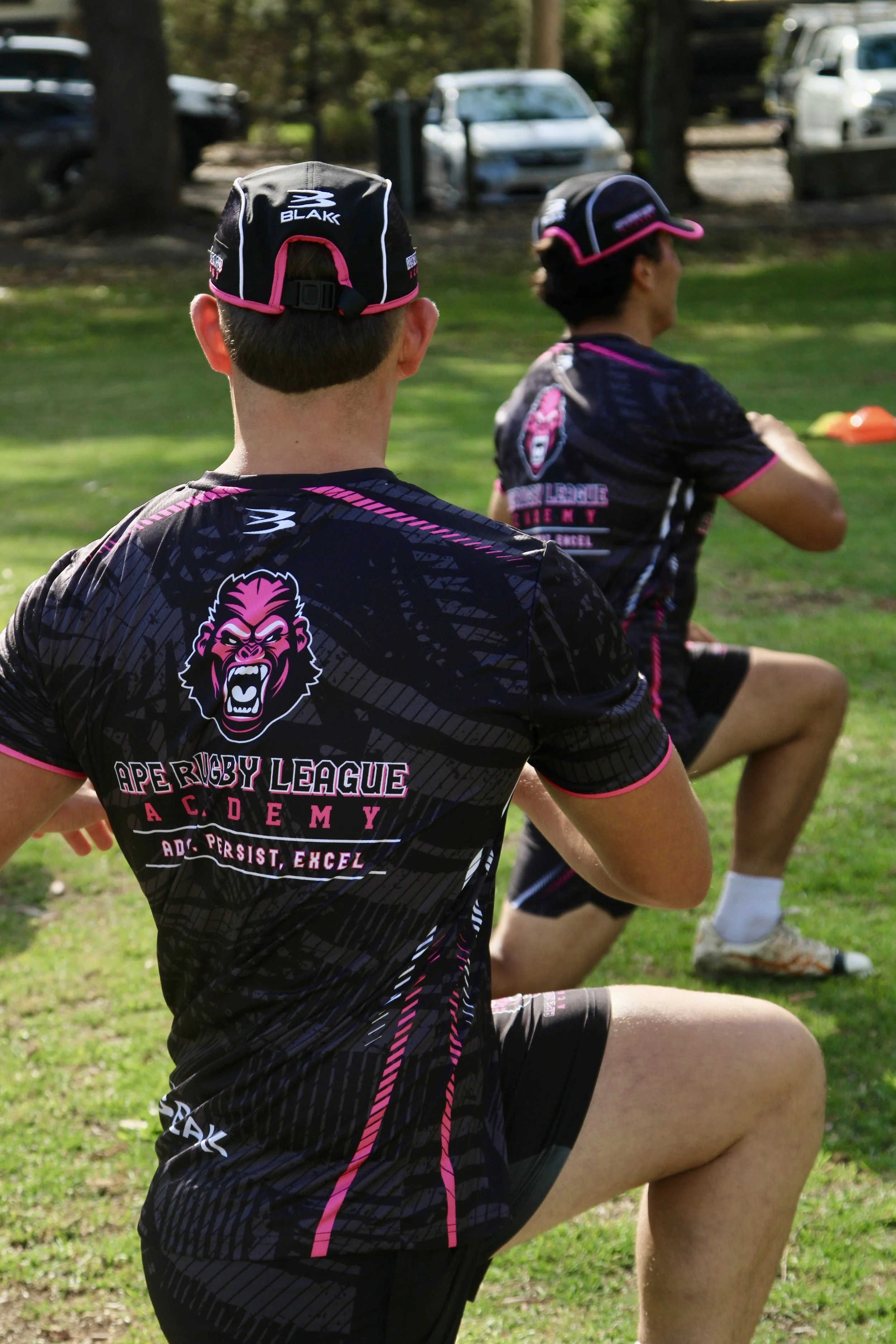 Two male rugby players wearing black and pink jerseys with a lion logo, sitting on grass during practice or warm-up.