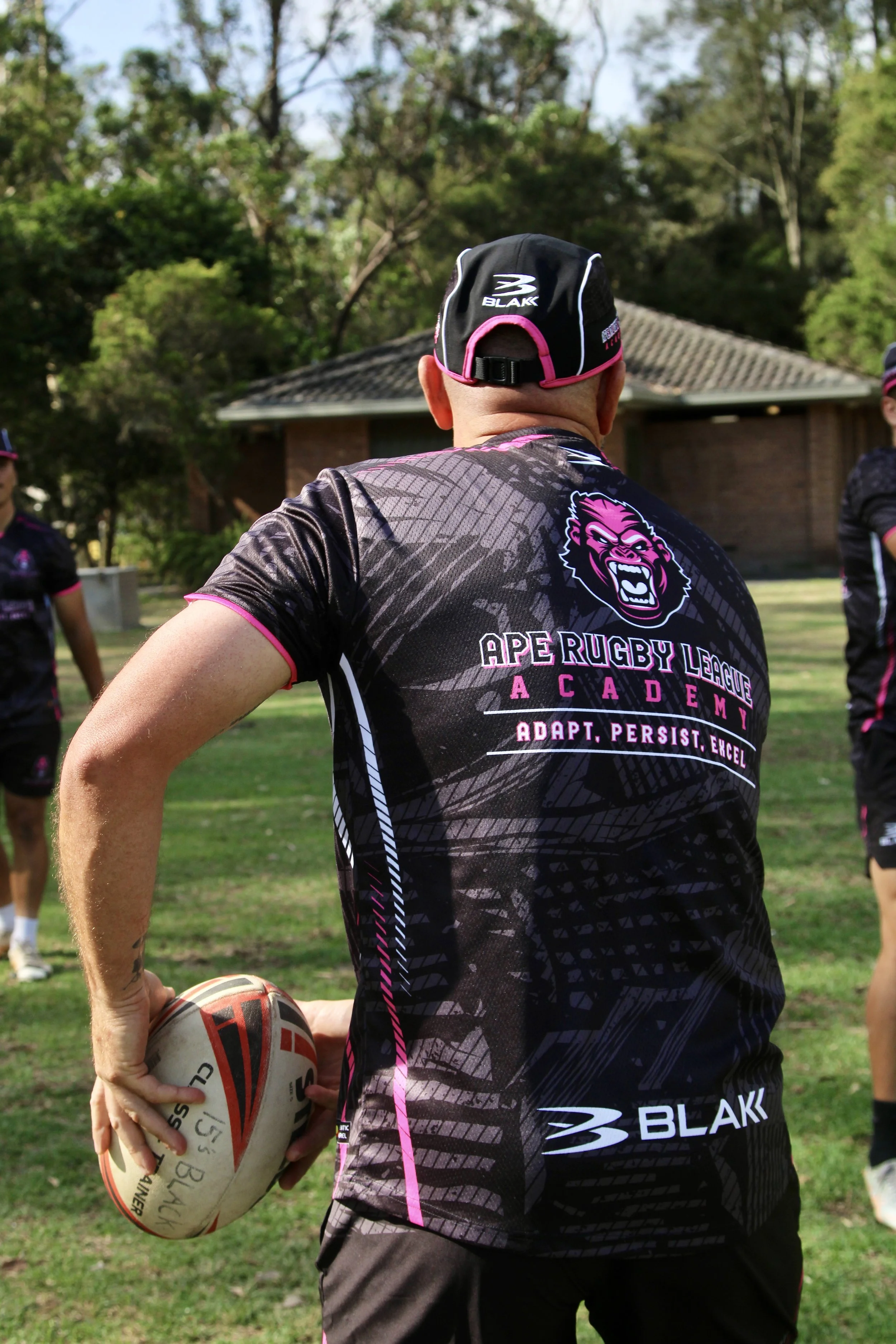 Person in rugby gear holding a rugby ball, standing outdoors on a grassy field with trees and a building in the background, participating in a rugby training session.