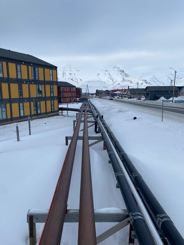 Strade innevate con tubi metallici che attraversano un paesaggio montano con edifici colorati e montagne innevate sullo sfondo, Longyearbyen, Isole Svalbard.