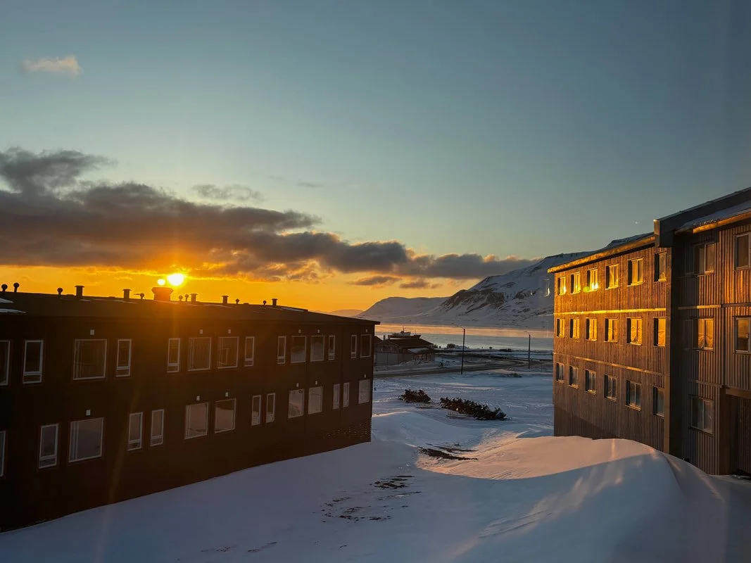Scorcio diurna di una zona urbana con edifici di legno circondata da neve, montagne sullo sfondo e il sole che tramonta all'orizzonte, Longyearbyen, Isole Svalbard.
