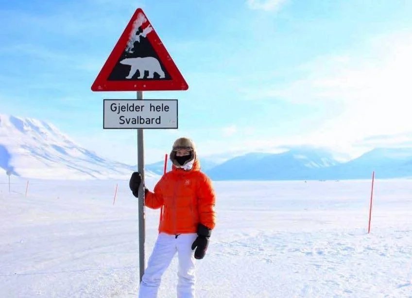 Persona che tiene un cartello con il segnale di attenti all'orso in un paesaggio innevato e montuoso, con testo in norvegese alle Isole Svalbard.