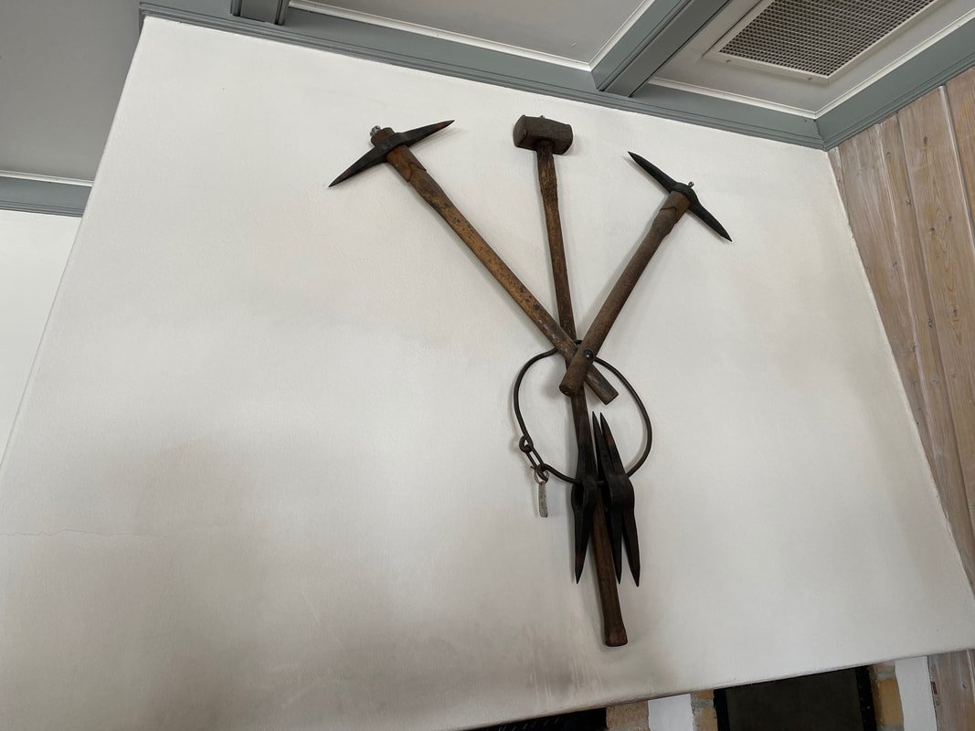 Un insieme di attrezzi di ferramenta appesi al muro, tra cui un piccone, un martello e due chiodi o scalpelli, disposti in modo decorativo.