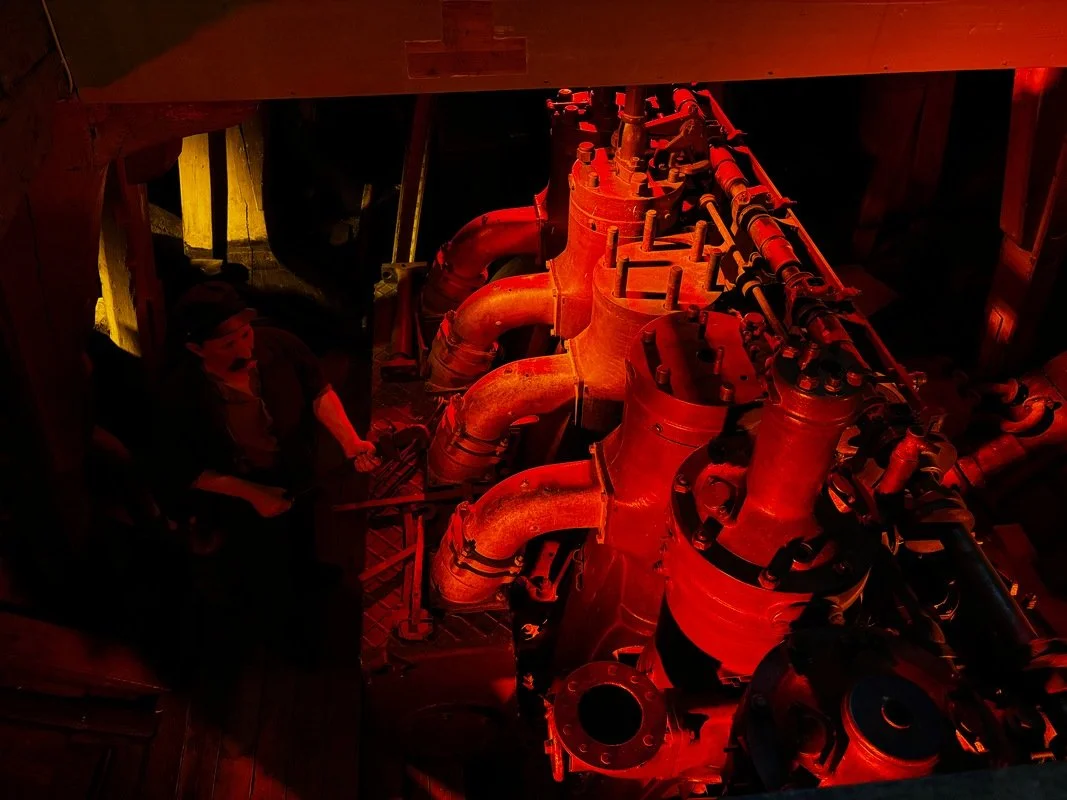 Immagine di una stanza con grandi macchinari industriali, con tubi e componenti metallici illuminati da luci rosse e gialle. Presente anche una figura umana che lavora o osserva.