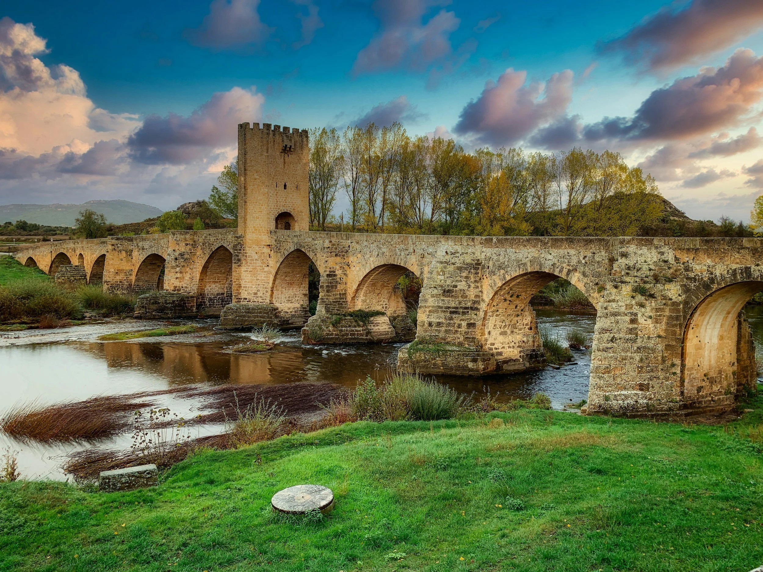 Un antico ponte in pietra con torres e archi attraversa un fiume, circondato da vegetazione e al tramonto, con un cielo ricco di nuvole.