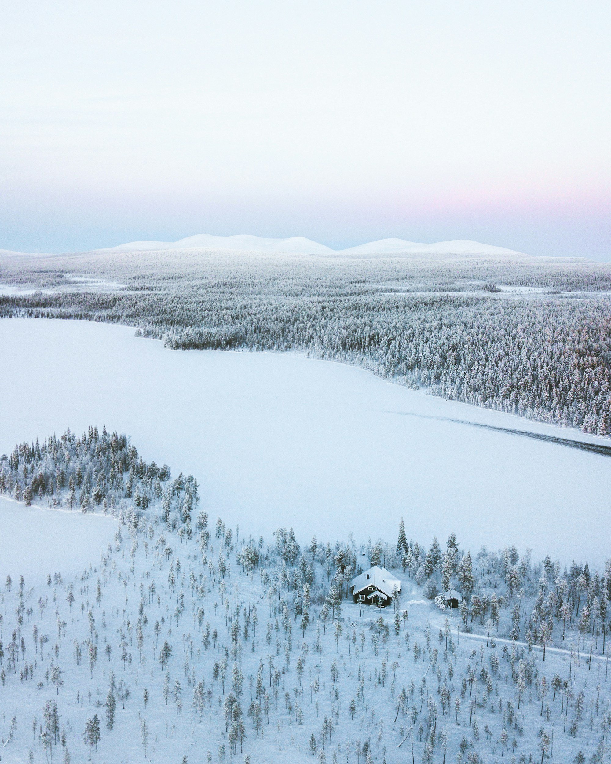 Paesaggio invernale con foresta di alberi ricoperti di neve, un lago ghiacciato e alcune case isolate, sullo sfondo montagne innevate sotto un cielo chiaro.