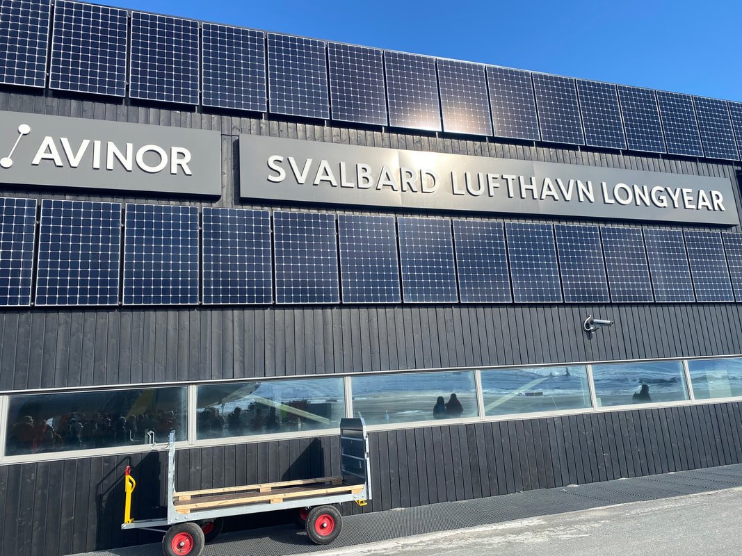 Facciata di un edificio con pannelli solari, segnale con scritte 'AVINOR SVALBARD LUFTHAVN LONGYEAR', finestra lunga e piattaforma di trasporto davanti.