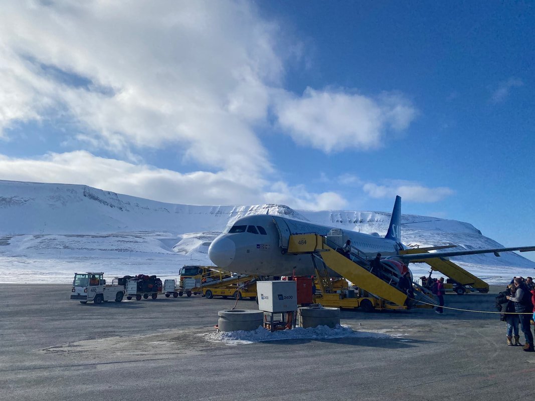 Aereo in fase di sbarco con scale e veicoli di supporto su una pista innevata di Longyearbyen alle Isole Svalbard, con montagne innevate sullo sfondo e cielo blu con nuvole.