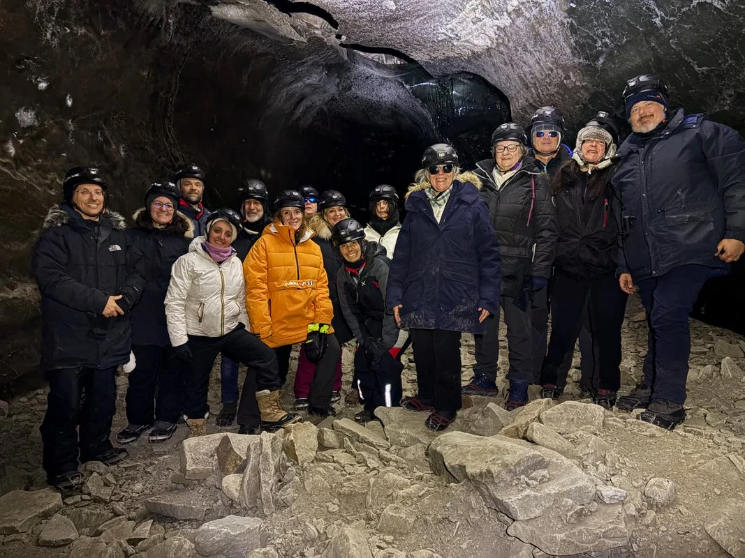 Gruppo di persone con caschi in una grotta o caverna, tutti vestiti con abbigliamento invernale e scarpe robuste, alcuni con occhiali da sole, in un ambiente roccioso e buio.