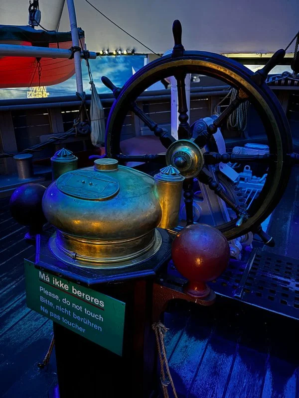 Antico timone della nave Fram con vari oggetti in metallo, una sfera rossa e un cartello con istruzioni multilingue