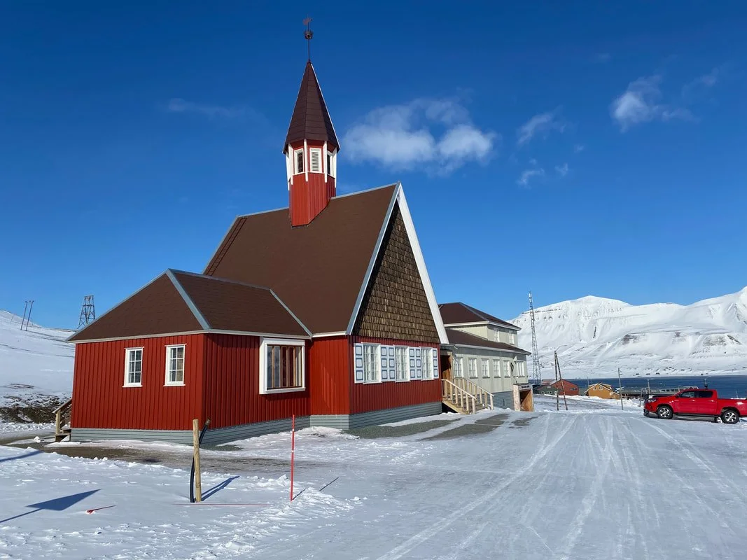 Chiesa di legno rossa a Longyearbyen, Isole Svalbard, con tetto ripido e campanile, circondata da neve e paesaggi montani invernali, con un'auto rossa parcheggiata e un cielo blu.