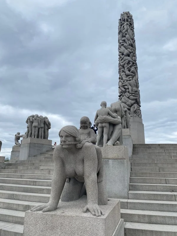 Sculture di pietra raffiguranti figure umane e un monolito decorativo, posizionate su gradini di pietra, sotto un cielo nuvoloso, parco Vigeland a Oslo.