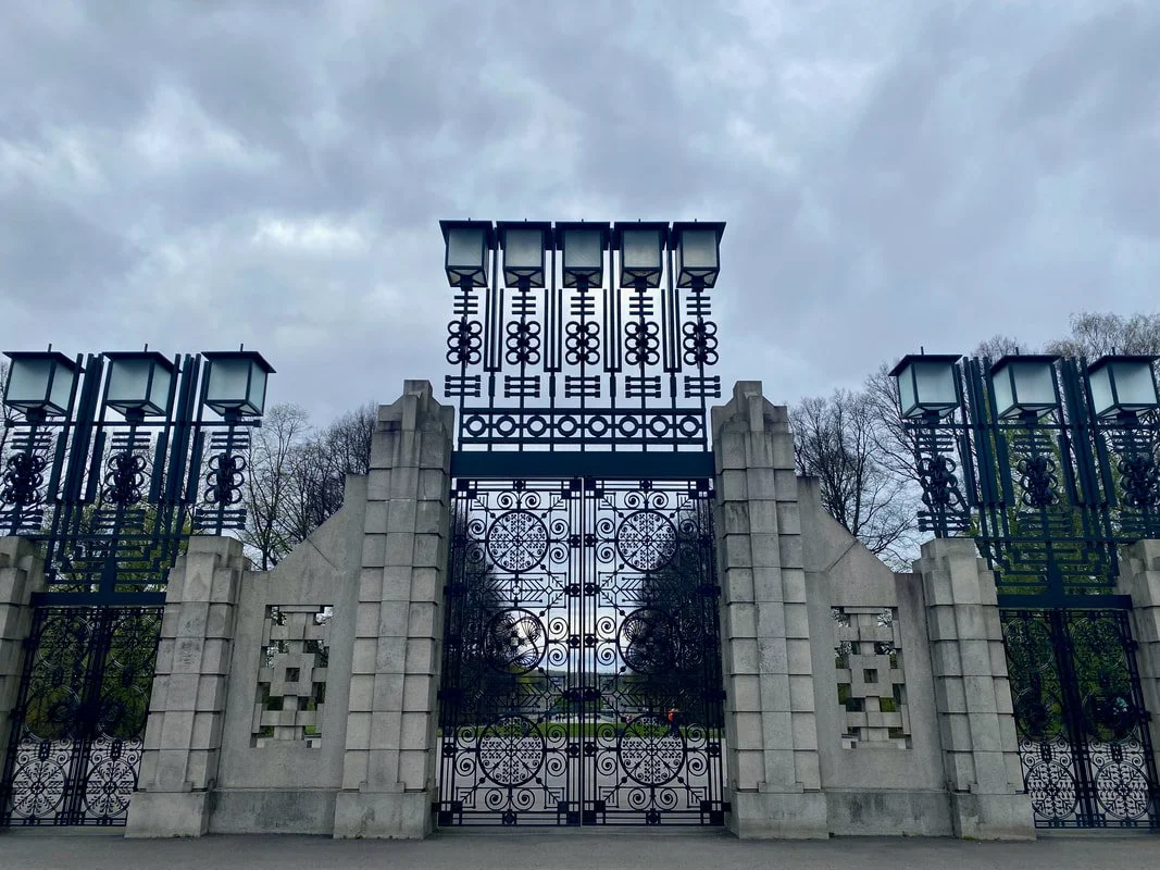Cancello di ferro nero decorato con motivi circolari del parco Vigeland a Oslo, con colonne di pietra su entrambi i lati, e lampioni sopra il cancello, sotto un cielo nuvoloso, con alberi spogli sullo sfondo.