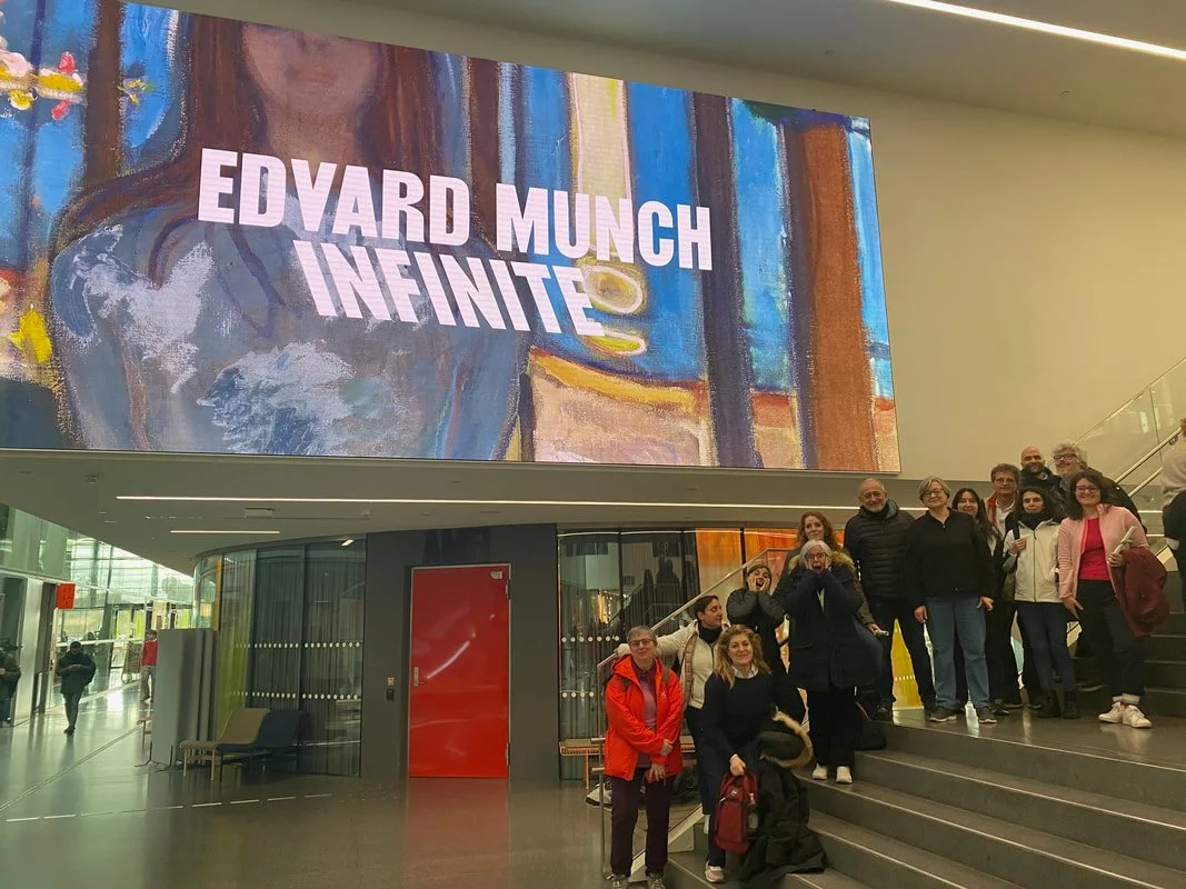 Gruppo di persone in visita a un'installazione artistica, con un grande schermo che visualizza il titolo 'EDVARD MUNCH INFINITE' in uno spazio moderno.