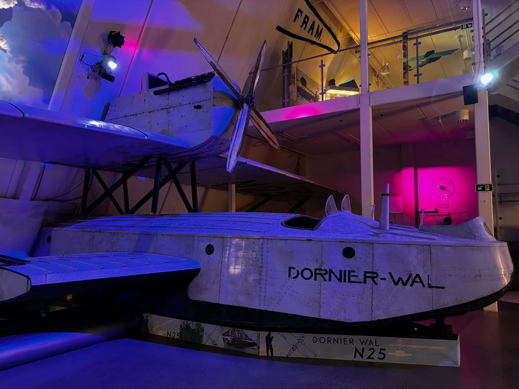 Una replica di un seaplane Dornier Wal esposta in un museo. L'aereo ha un design bianco con il nome 'DORNIER-WAL' e 'N25' sulla fiancata. È esposto in un ambiente con luci colorate, tra cui luci blu, viola e rosa.