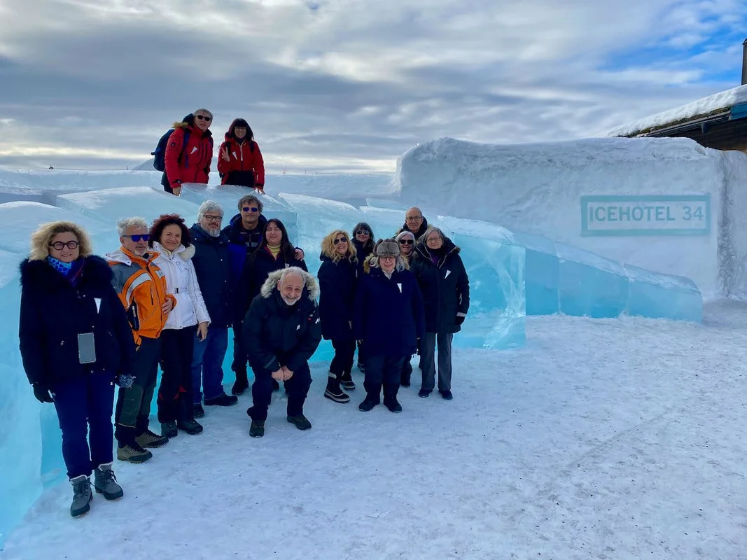 Gruppo di persone davanti all'Ice Hotel di Jukkasjärvi in Svezia, con un cartello che recita ICEHOTEL 34, in un paesaggio innevato con cielo nuvoloso.