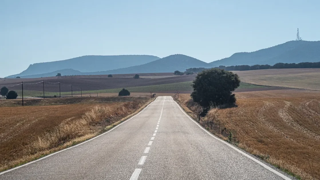 Una strada di campagna che si estende verso le colline sullo sfondo, con alcuni alberi e campi circostanti.