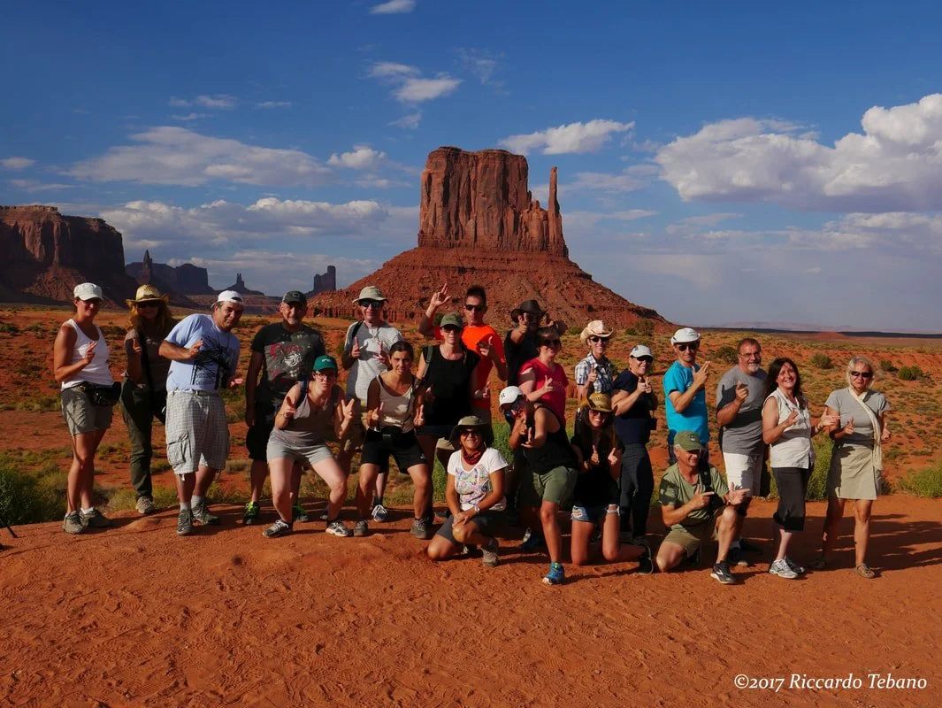 Gruppo di persone davanti a monumenti naturali nel deserto della Monument Valley negli Stati Uniti d'America sotto un cielo blu, alcune in posa, altre parlando o scattando foto.