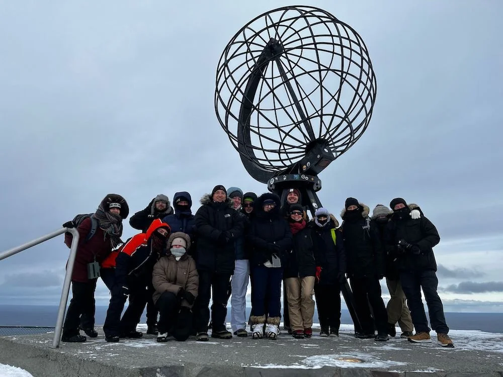 Gruppo di persone sulla sommità di una piattaforma con la grande sfera di metallo di Capo Nord, in mezzo a un paesaggio nevoso con cielo nuvoloso.