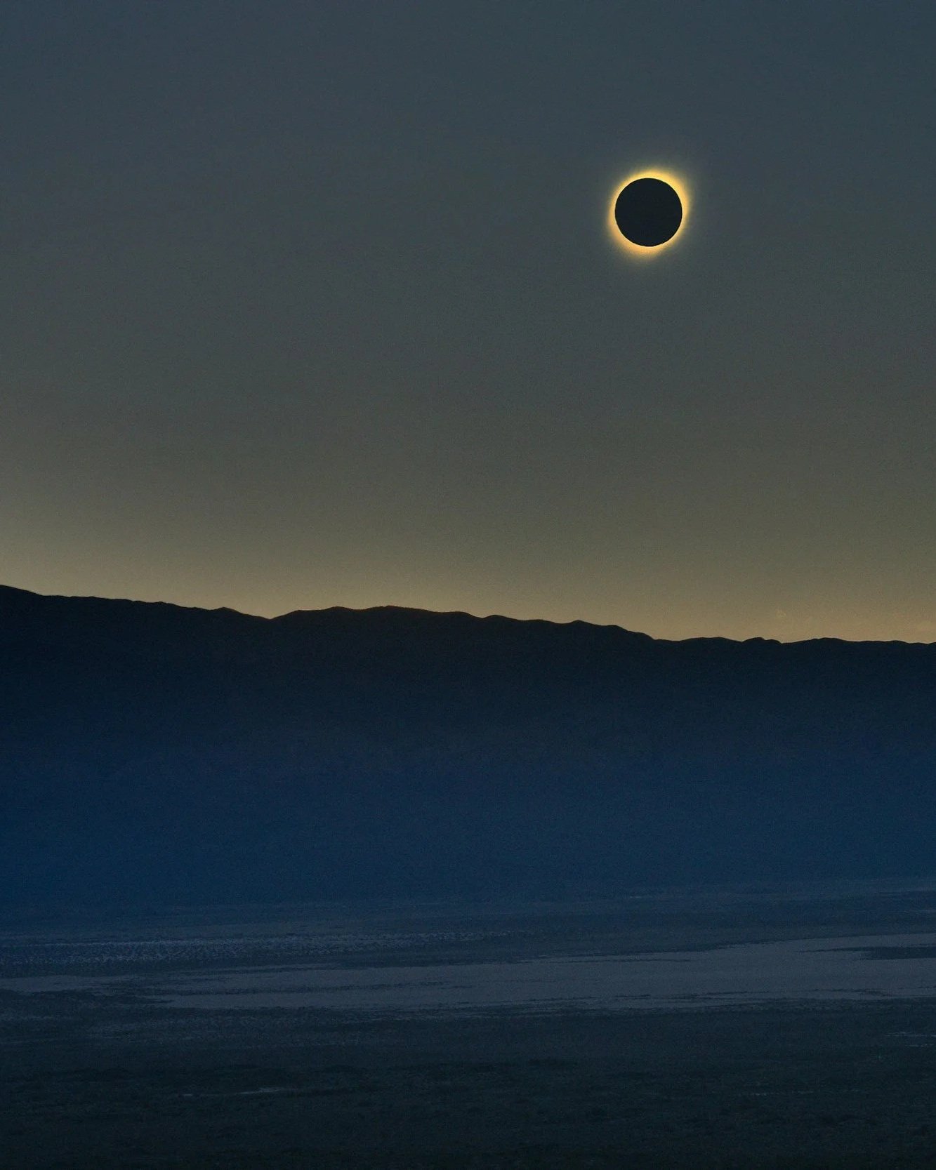 Eclissi solare con il cielo scuro e la luna che copre il sole, vista da una spiaggia con silhouette di montagne sullo sfondo.