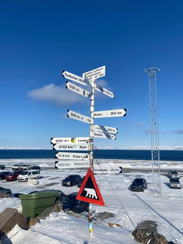 Pali in metallo con segnali di destinazione e distanza in chilometri, all'esterno dell'aeroporto di Longyearbyen Svalbard, in riva al mare ghiacciato con alcune vetture parcheggiate e un cielo sereno.