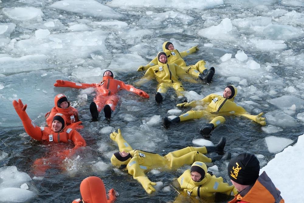 Persone in tute di immersione rosa e gialle, alcune sdraiate e altre galleggianti nell'acqua di un ambiente ghiacciato con pezzi di ghiaccio galleggianti.