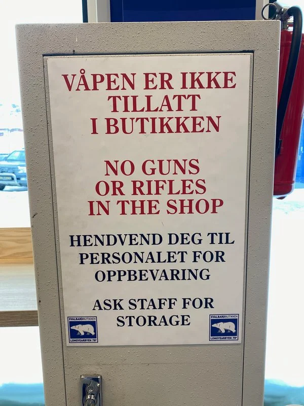 Cartellone con avviso scritto in norvegese e inglese che indica che non sono ammesse armi nel negozio, e invita a chiedere allo staff per lo stoccaggio, Longyearbyen, Isole Svalbard.