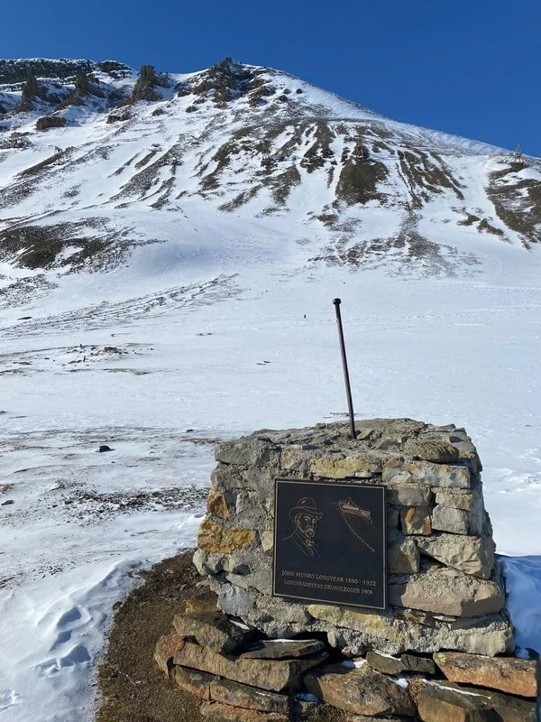 Pannello commemorativo sulla neve, con montagne innevate sullo sfondo, e un cielo blu, a Longyearbyen, Isole Svalbard.