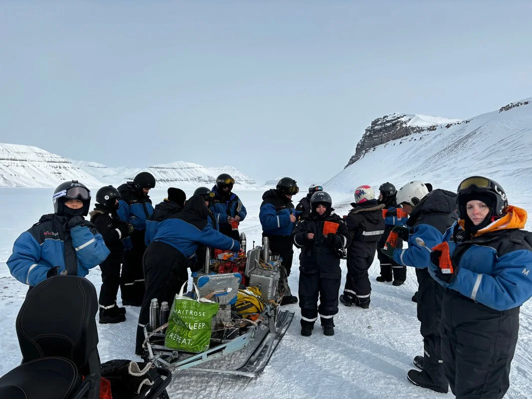 Gruppo di persone con abbigliamento da sci in una zona di neve tra montagne, alcune con caschi e occhiali protettivi, intorno a un'attrezzatura per attività invernali, Tempelfjörd, Isole Svalbard.