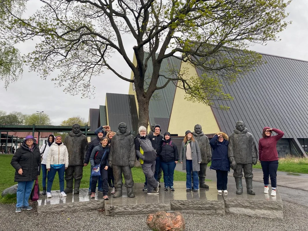 Gruppo di persone che posano davanti a statue di bronzo di diverse figure umane al museo Fram di Oslo, sotto un albero con foglie verdi.