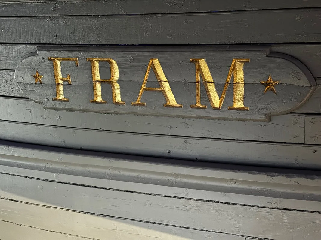 Scritta decorativa con la parola 'FRAM' in lettere dorate su sfondo di legno grigio.