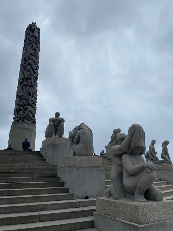 Sculture di figure umane in pietra su una piattaforma con gradini, con un monumento con rovine intrecciate sullo sfondo e un cielo nuvoloso sopra, parco Vigeland a Oslo.