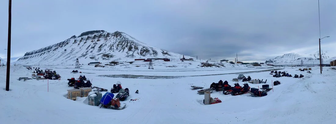 L'immagine mostra un paesaggio innevato con motoslitte parcheggiate, un monte innevato sullo sfondo e alcune strutture sparse nel panorama, Longyearbyen, Isole Svalbard.