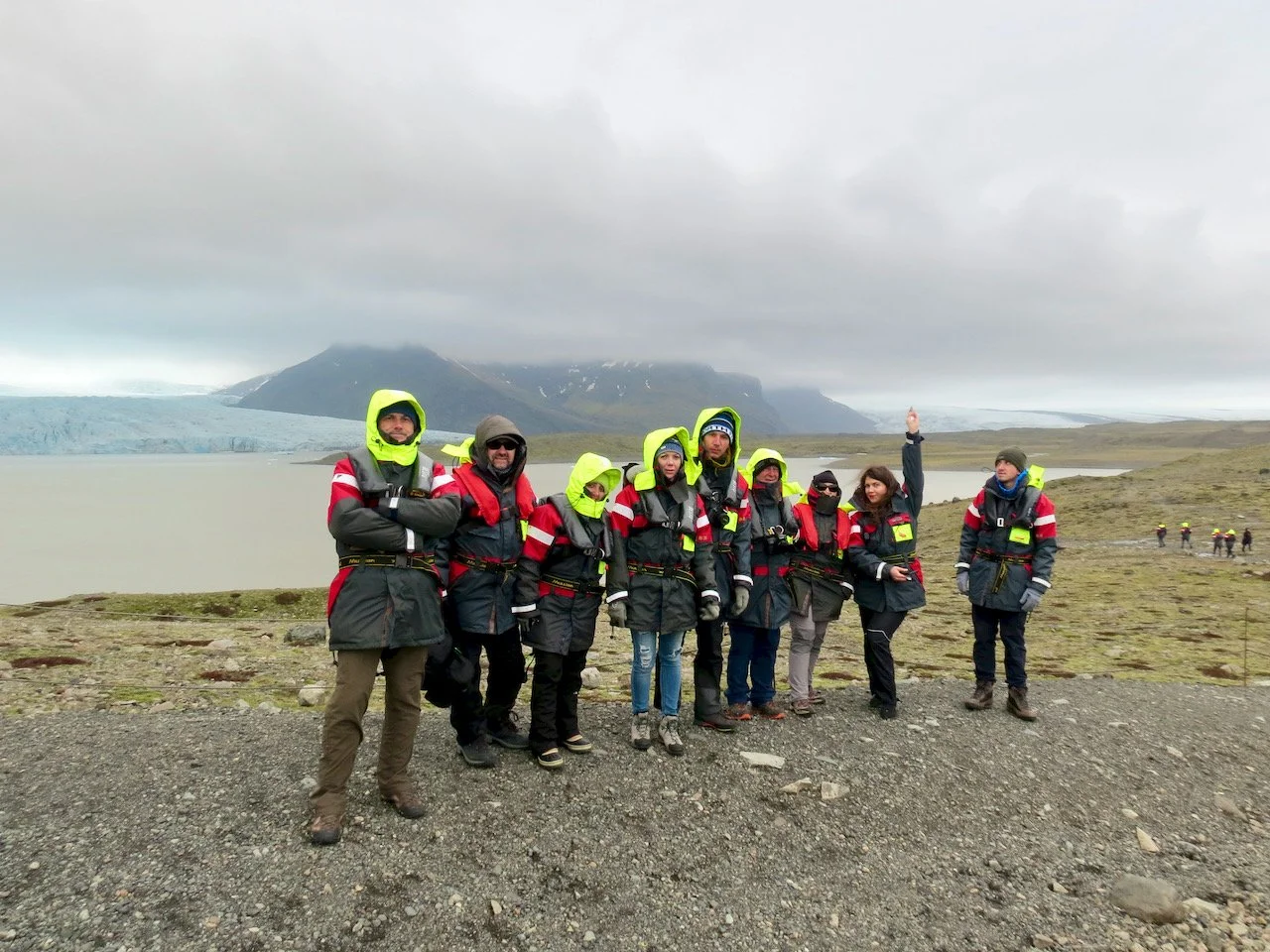 Gruppo di persone in abbigliamento da escursione, con cappucci gialli e giacche nere con strisce rosse, in una zona vulcanica con un ghiacciaio islandese sullo sfondo.