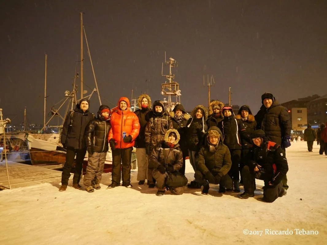 Gruppo di persone con abbigliamento invernale in una banchina di porto a Trömso con barche e imbarcazioni sullo sfondo, sotto un cielo notturno piovoso.
