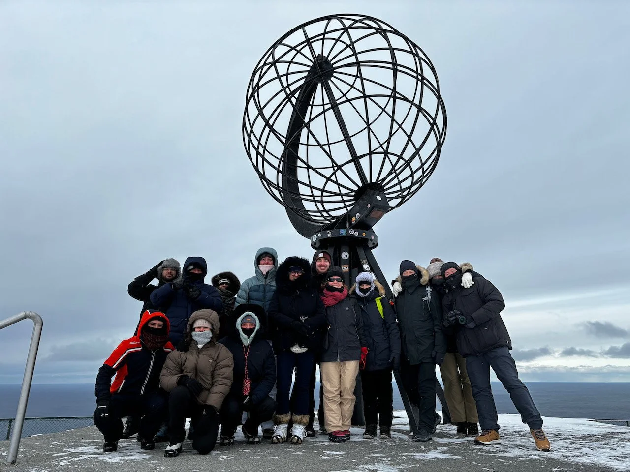 Un gruppo di persone in abbigliamento invernale davanti alla grande sfera metallica di Capo Nord, con paesaggio marino sullo sfondo