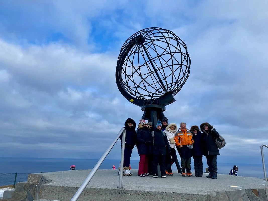 Gruppo di persone in piedi vicino al grande globo di metallo di Capo Nord, con il mare sullo sfondo e nuvole nel cielo.