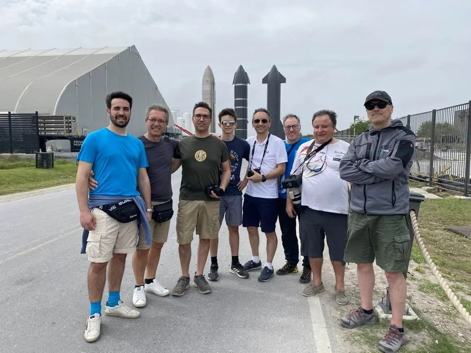 Un gruppo di otto persone sorridenti posa per una foto all'aperto nella base SpaceX a Boca Chica in Texas con sullo sfondo tre razzi Starship.