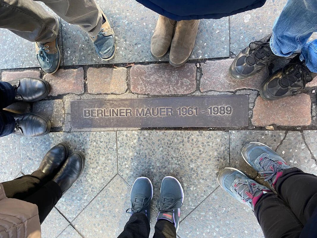 Piazza con mattonelle e una piastra di metallo con scritto "Berliner Mauer 1961-1989" circondata da piedi di persone con scarpe diverse.