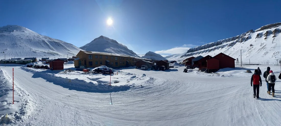 Paesaggio invernale con case rosse, montagne innevate e persone che camminano sulla neve sotto un cielo sereno con il sole, Longyearbyen, Isole Svalbard.
