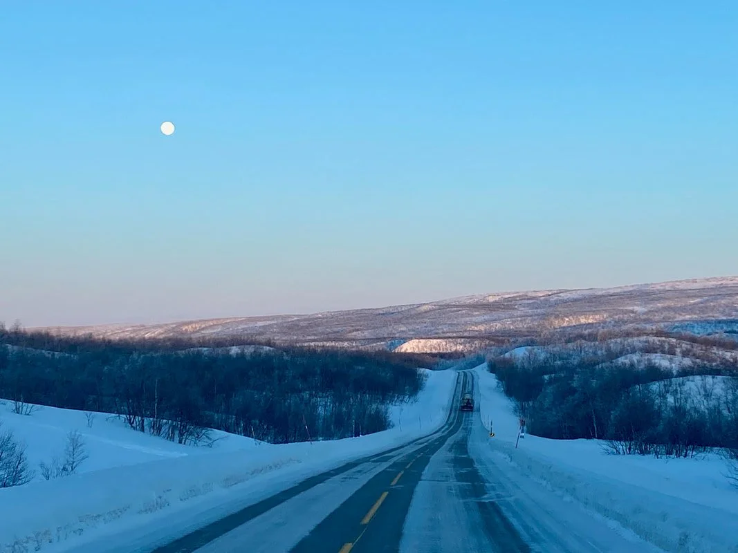 Strada innevata che si snoda tra le colline ghiacciate sotto un cielo azzurro con la luna visibile nella Lapponia finlandese.